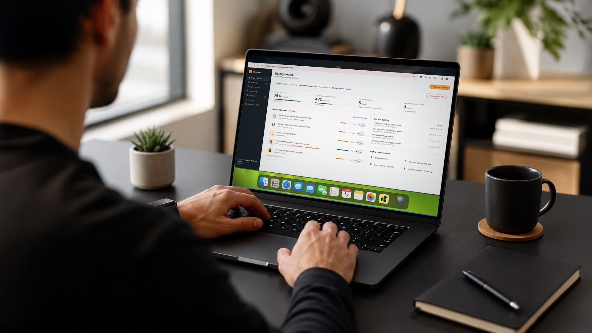 A designer at a laptop on a dark desk, the screen showing the AI DAM Librarian dashboard with library health scores, a review queue, and recent agent activity