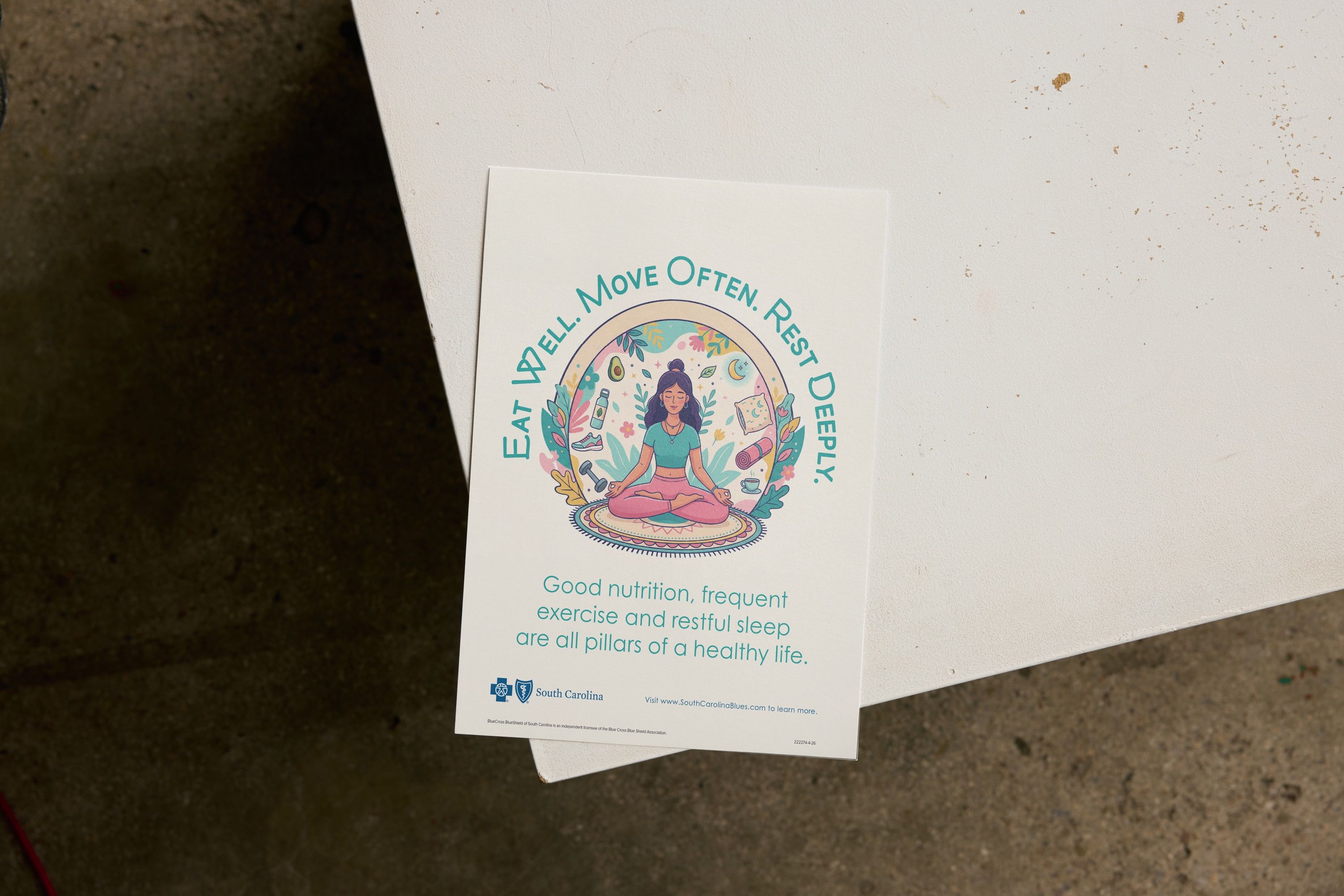 The "Eat Well. Move Often. Rest Deeply." flyer lying on a white table: a woman meditates inside a circle of healthy-living icons under an arched headline