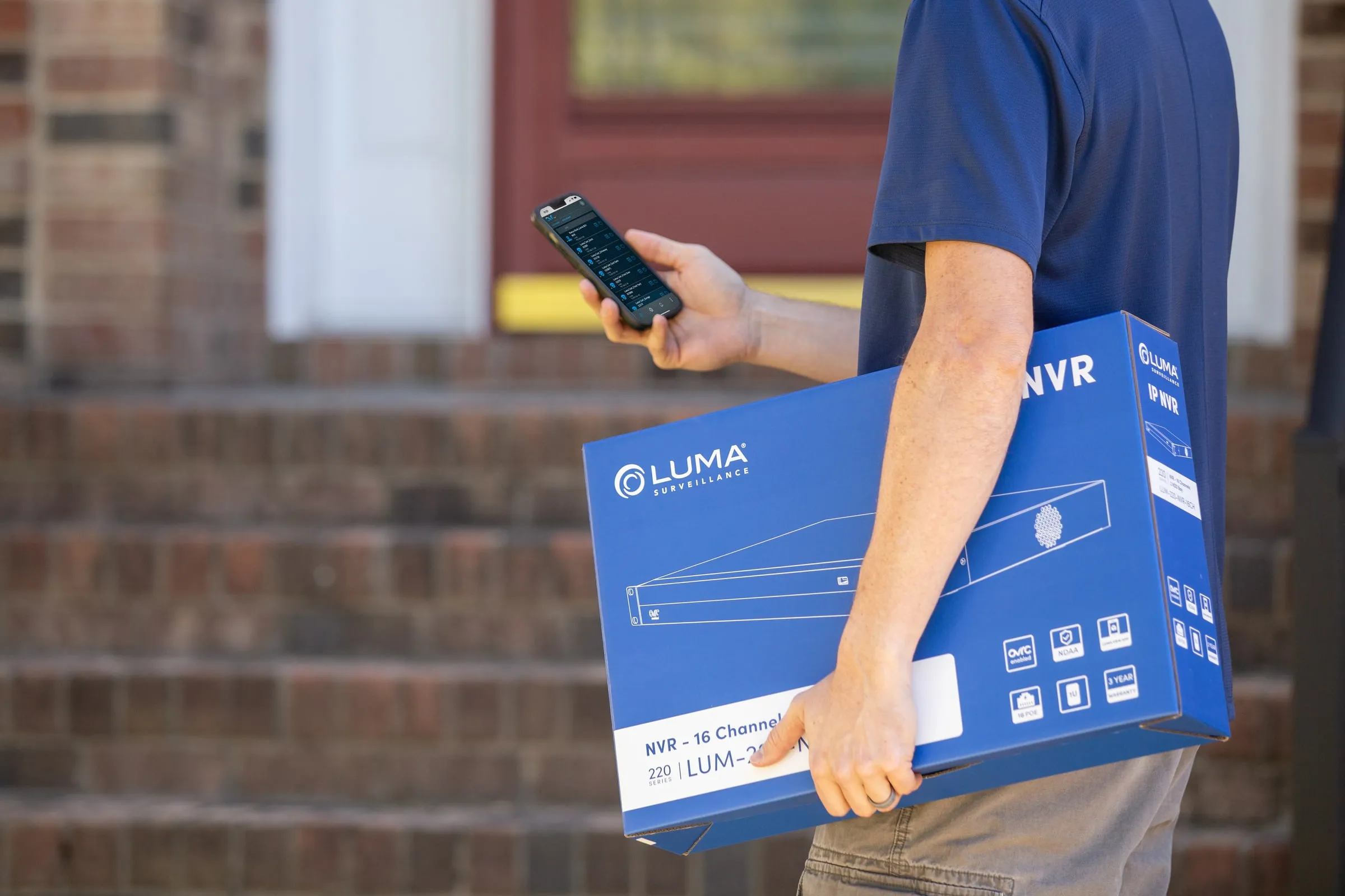 Person carrying a blue Luma NVR box while checking a phone