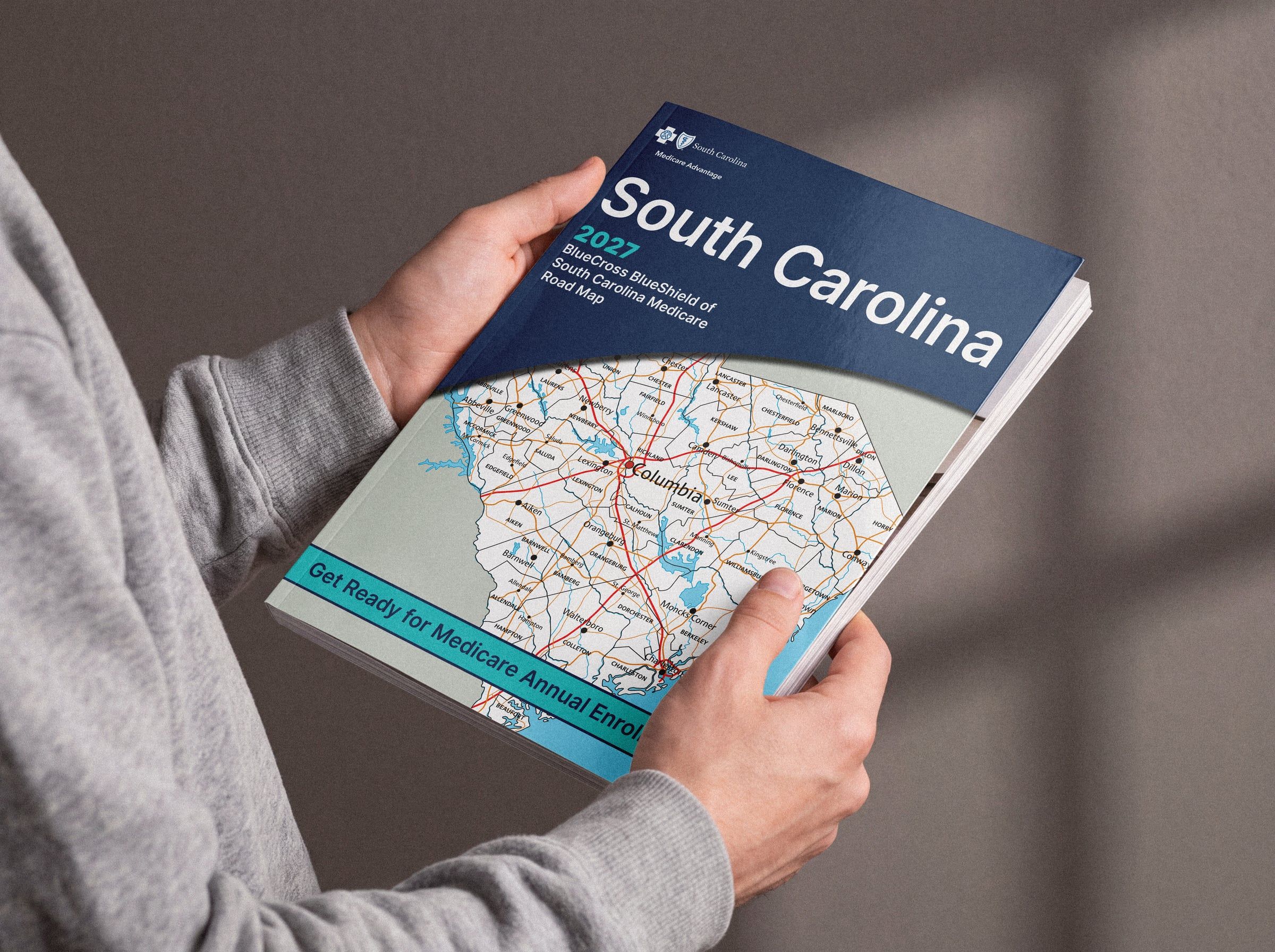 Two hands hold the printed 2027 BlueCross BlueShield of South Carolina Medicare Road Map. The cover is a road map of South Carolina under a navy band with the title “South Carolina” and a teal strip reading “Get Ready for Medicare Annual Enrollment!”