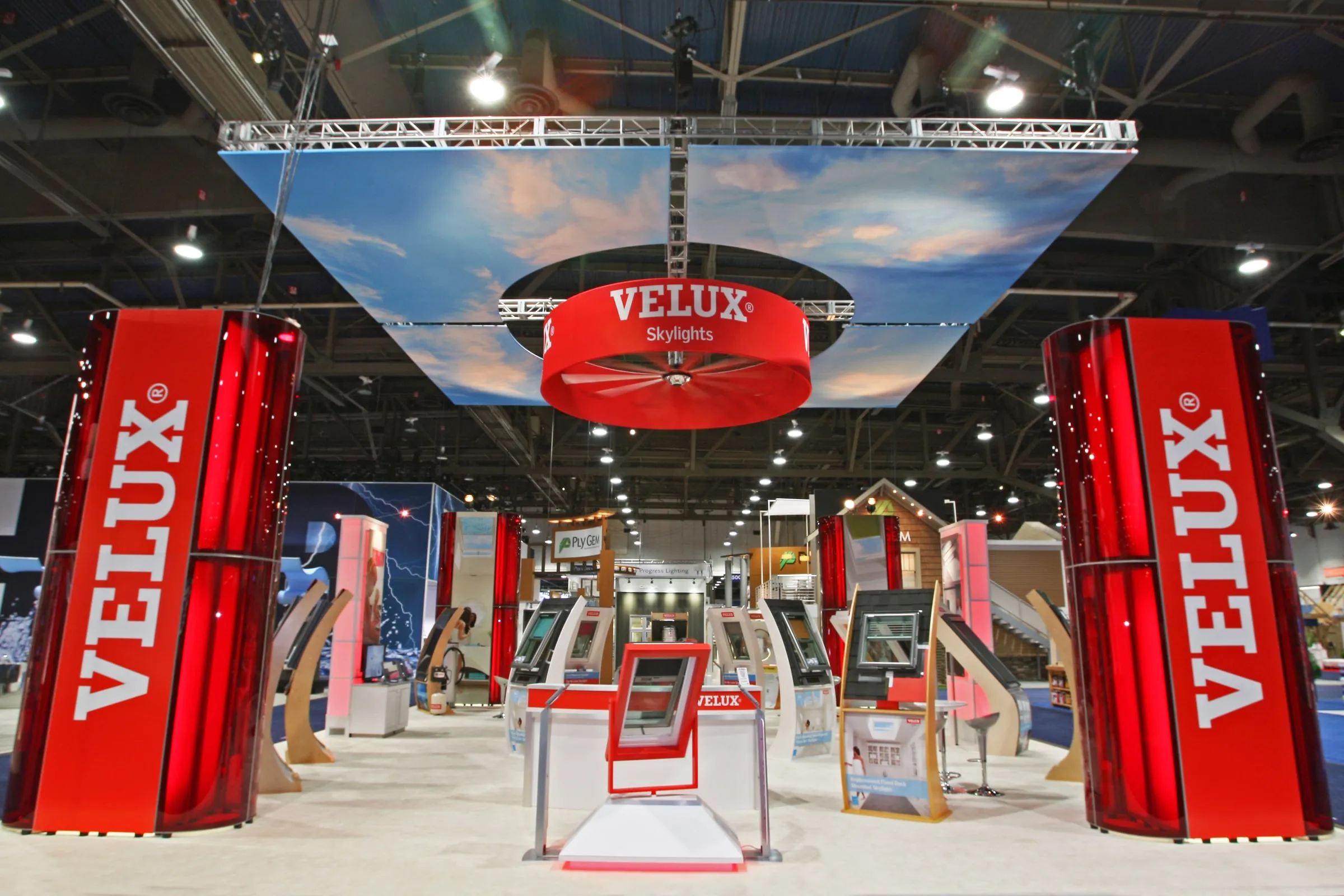 Wide view of a VELUX trade show booth with tall red VELUX towers and an overhead sky banner (1 of 3)