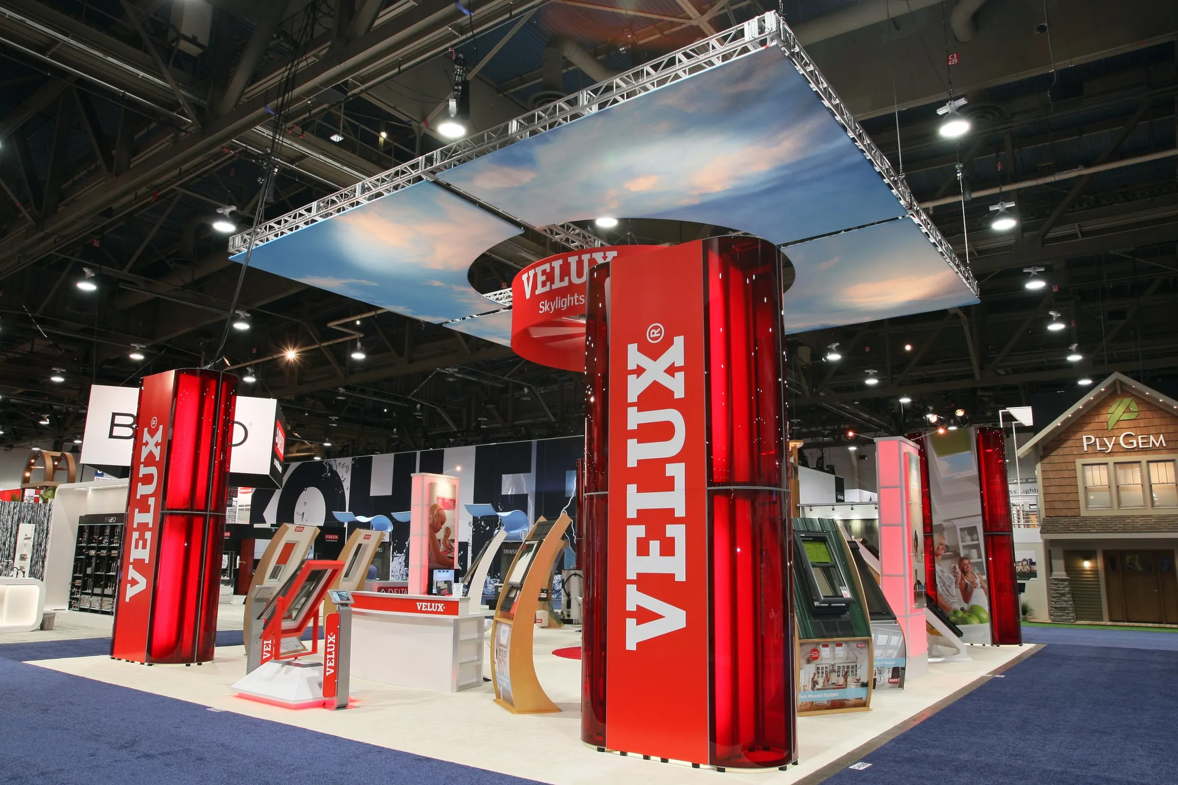Wide view of a VELUX trade show booth with tall red VELUX towers and an overhead sky banner (2 of 3)